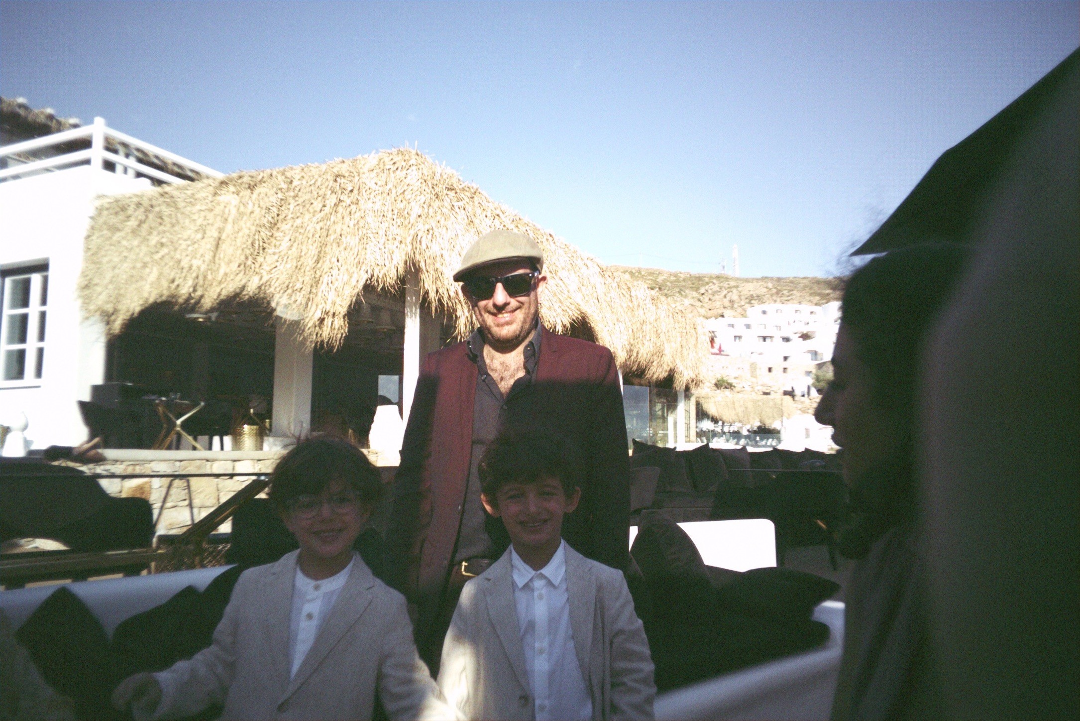 Remi wearing sunglasses and a flat cap, standing with two kids in front of a thatched-roof beach bar
